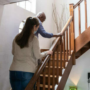 Home caregiver helping senior man walking on the staris at home - wearing face mask